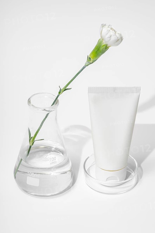 A modern skin care laboratory setup with a white flower stem extending through a clear flask and a white tube of cream, symbolizing organic and scientific beauty product development