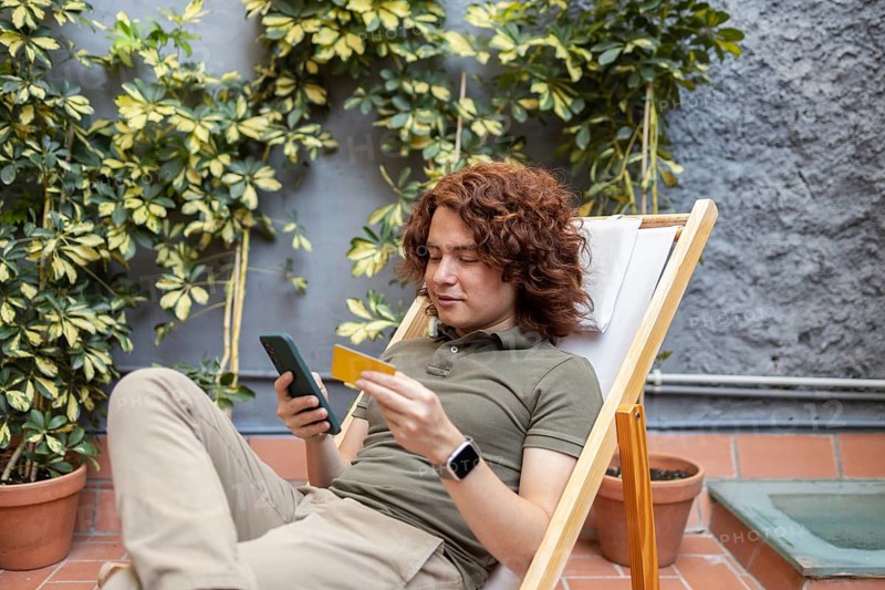 A young man lounging outdoors in a chair, holding a smartphone and a credit card. He is casually dressed, enjoying a blend of relaxation and technology in nature