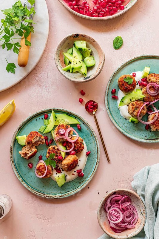 A vibrant Mediterranean dish featuring meatballs, fresh cucumbers, yogurt, pomegranate seeds, and red onions on a turquoise plate. Styled with herbs and a subtle background