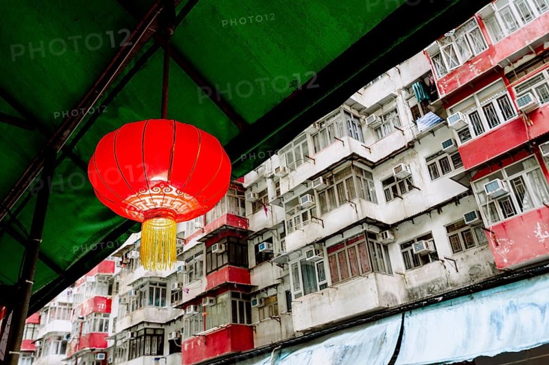 A bright red Chinese lantern hangs under a green awning, set against the densely packed, weathered apartment buildings in Hong Kong, celebrating Chinese New Year
