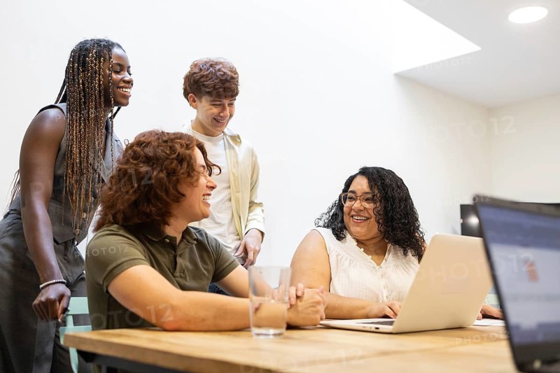A diverse group of colleagues collaborate in a modern office setting. They are engaged in a positive discussion around a table with laptops, fostering an inclusive work environment