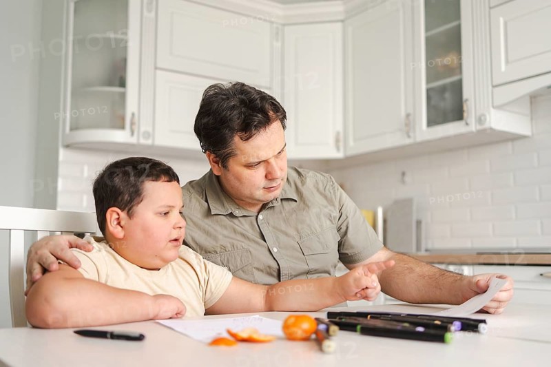 A father helps his son with homework at the kitchen table. They are surrounded by school supplies, fostering a learning environment and family bonding
