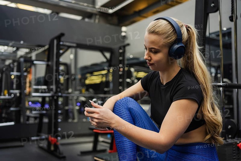 A blonde woman with headphones relaxes while checking her phone at a gym. She wears fitness attire and is surrounded by exercise equipment, creating an active atmosphere