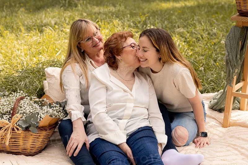 A warm and joyful moment during a family picnic features a grandmother, mother, and daughter seated on a blanket in a park. They share laughter and smiles under the sunny sky