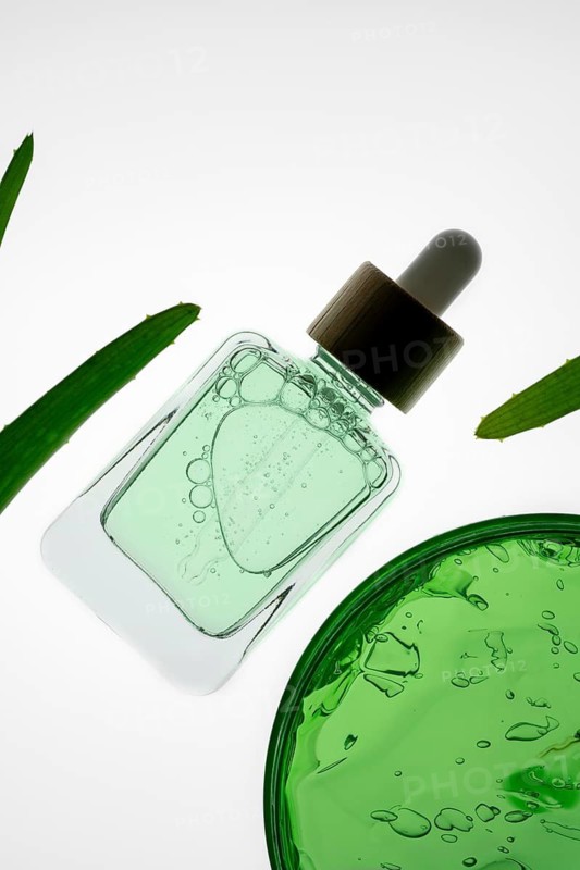 A bottle of aloe vera serum with a dropper lies on a white background, surrounded by green leaves. The minimalist arrangement evokes a sense of natural beauty and freshness