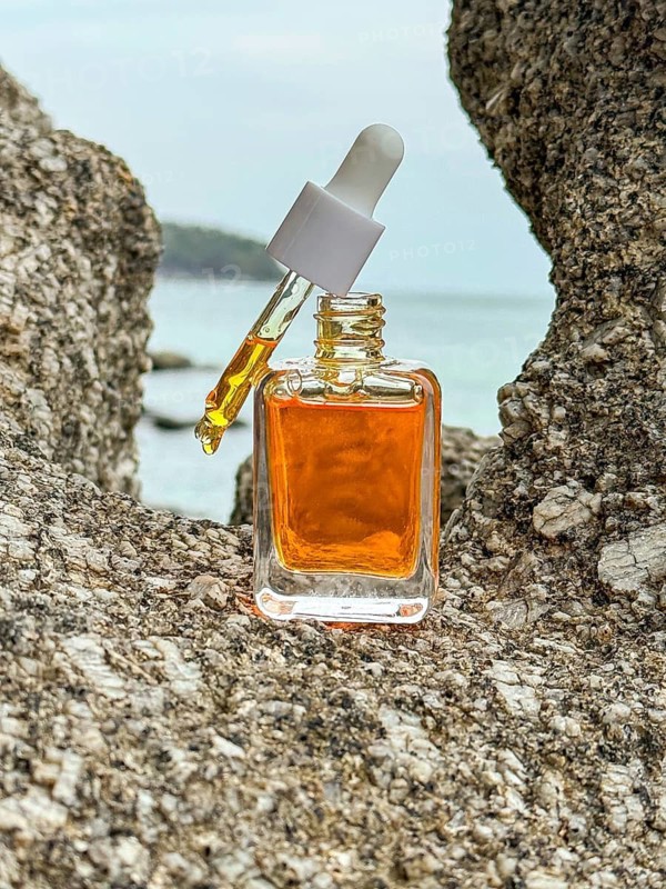 Between rugged rocks, a luxurious cosmetic serum bottle infused with vitamins and shimmering golden particles stands with the ocean backdrop, symbolizing high end natural skincare