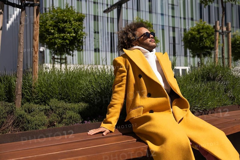 Afro man relaxing on a wooden bench outdoors, wearing a fashionable yellow coat and sunglasses, enjoying a sunny day in an urban green space, surrounded by plants