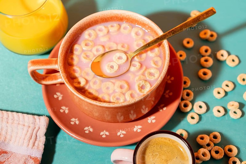 Top view of a colorful breakfast setting featuring a ceramic bowl of cereal with milk, a cup of coffee, and orange juice on a turquoise surface