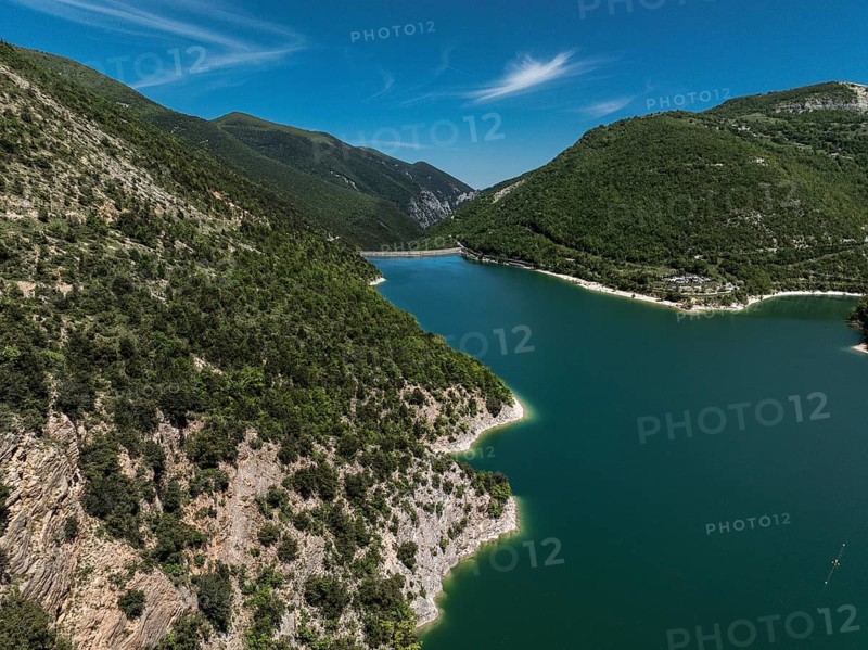 A captivating aerial view of Lago di Fiastra in Italy, surrounded by lush green hills and clear blue skies. The tranquil lake reflects the natural beauty of the landscape