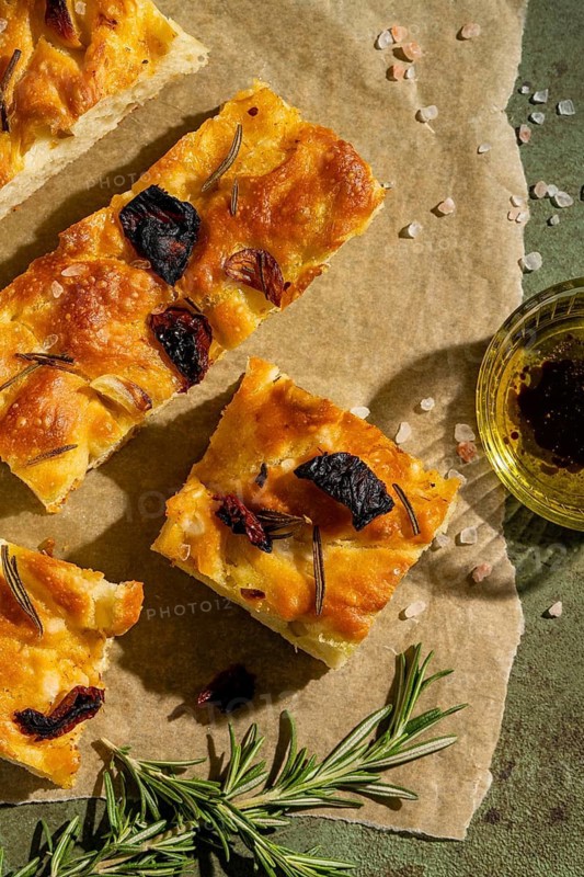 Rustic squares of homemade focaccia bread, topped with rosemary and sun dried tomatoes, rest on parchment paper. A side of olive oil dip completes this savory delight