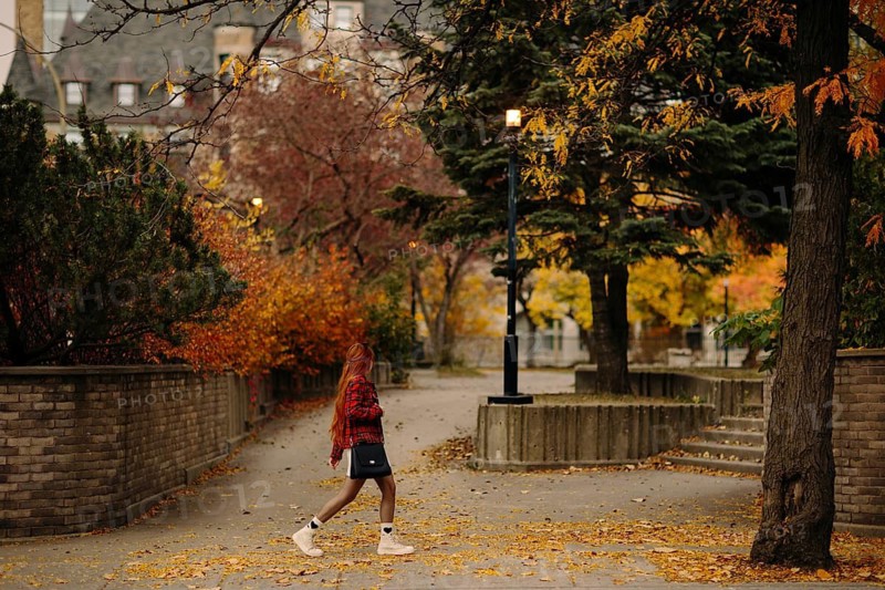 Side view of unrecognizable redhead woman walking through a vibrant autumn scene in Montreal. The pathway is lined with colorful trees and fallen leaves