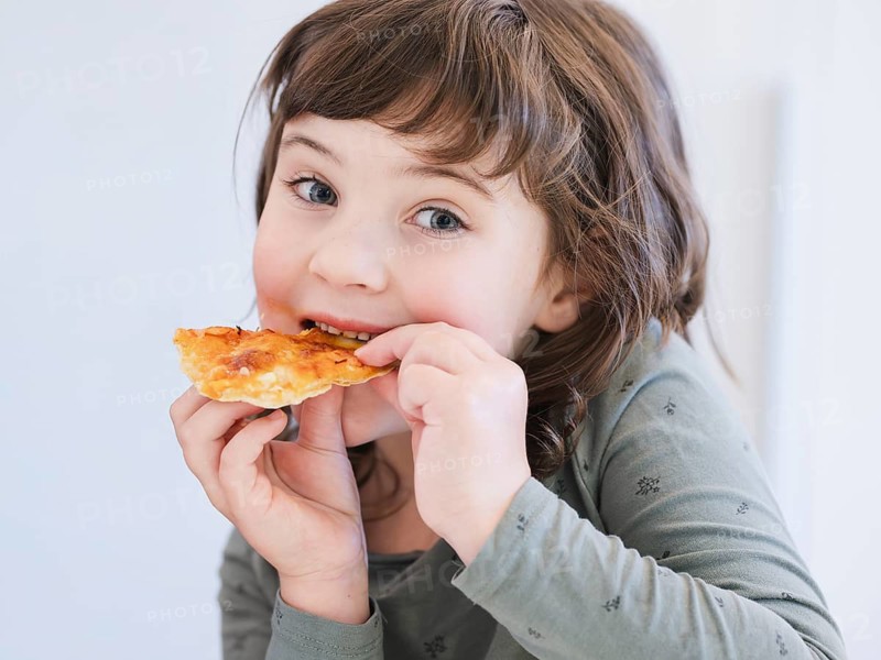 A young child joyfully eats a slice of pizza, showcasing the simple delights of childhood. The bright eyes and slight grin capture a moment of pure happiness