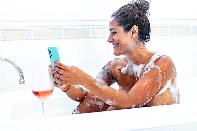 A woman relaxes in a bubble filled bath, using her smartphone with a smile. A glass of wine rests nearby, adding a touch of luxury to her serene moment at home