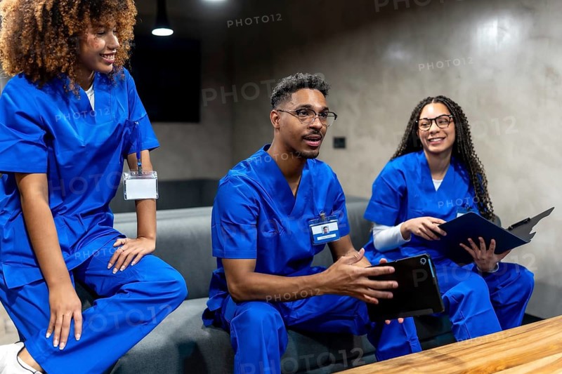 Group of diverse healthcare professionals in blue scrubs talking and sharing information while sitting in a modern common area, representing concepts of collaboration and ongoing learning