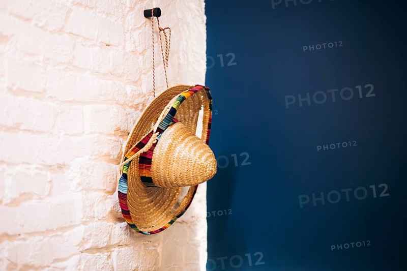 A traditional woven sombrero with colorful, vibrant trim hangs on a rustic white brick wall against a contrasting deep blue background, showcasing Mexican interior details