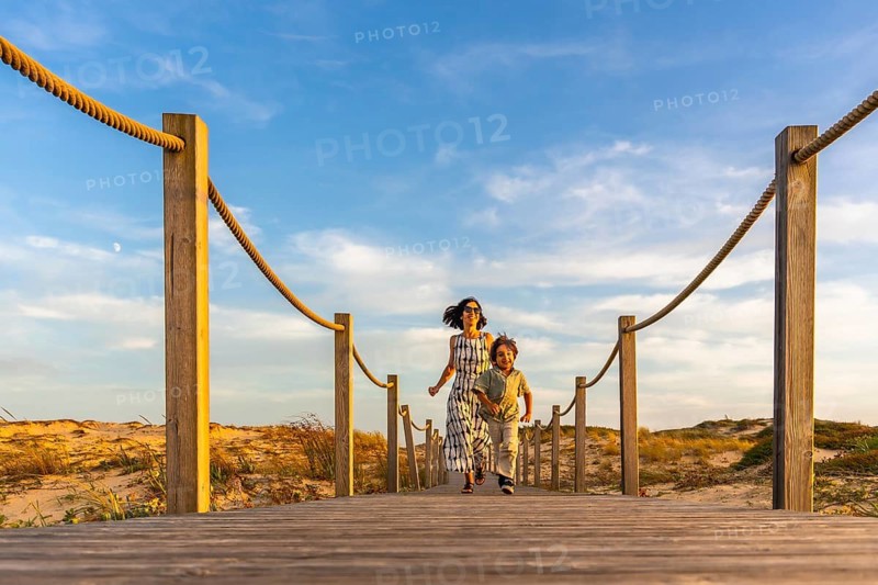 Happy mother and son running along a scenic wooden boardwalk leading to the beach, with sand dunes and golden light under a blue sky, enjoying family summer vacation in seignosse, france