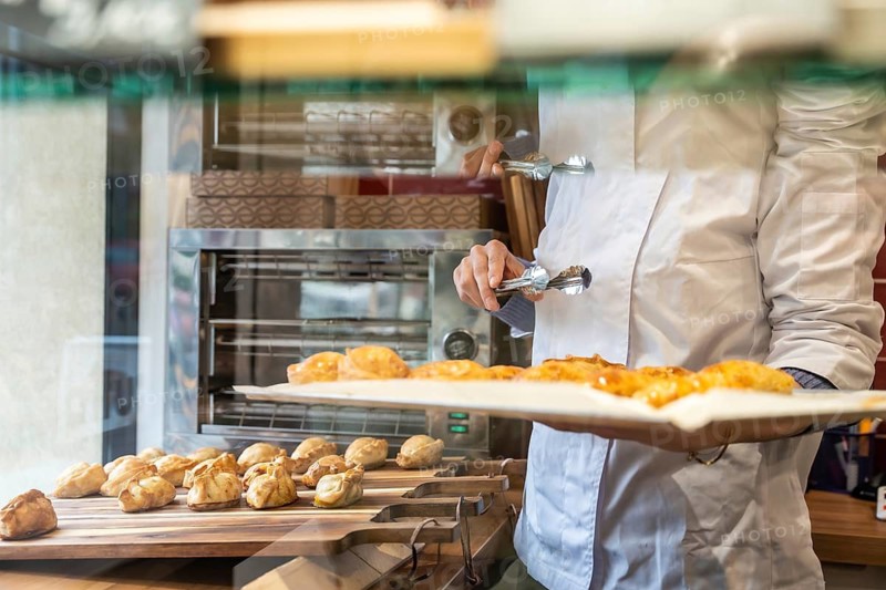 Chef wearing a white uniform holding a tray with freshly baked pastries, using tongs to move sweet empanadas and croissant like treats in a commercial bakery setting