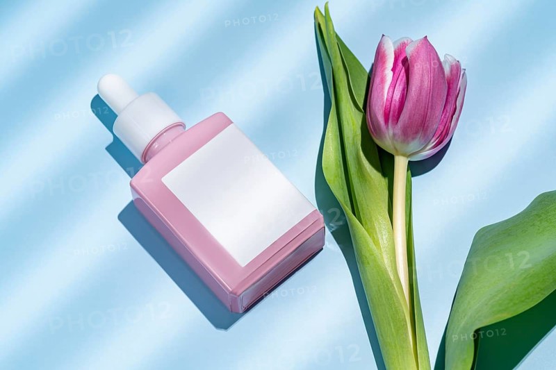 Top view of a pink cosmetic dropper bottle alongside a beautiful pink tulip, presented on a textured blue background. Ideal for beauty and skin care themes