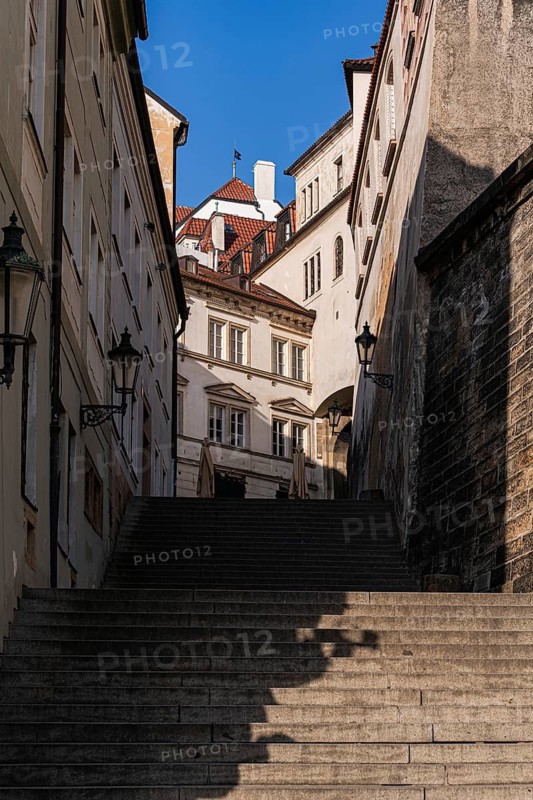 A narrow medieval street with steep stone stairs, surrounded by historic buildings with ornate windows and charming lanterns in Prague. Bright sunlight casts shadows on the pathway