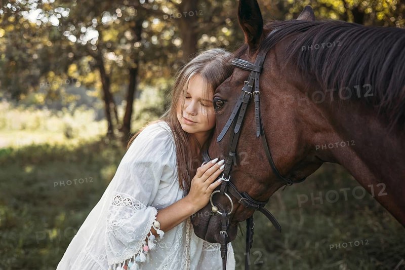 A young girl in a white dress gently embraces a brown horse, illustrating a serene bond in a sunlit forest setting. The scene reflects tranquility and connection