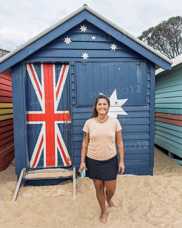 A cheerful tourist stands in front of a vibrant Brighton Beach hut in Melbourne, Australia. The iconic beach hut sports the vibrant colors of the Australian flag, creating a cheerful atmosphere