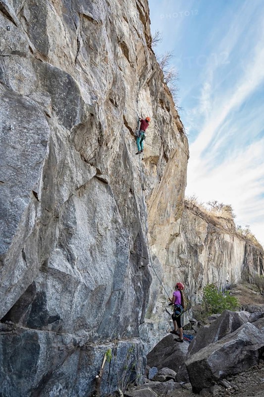 Two climbers face a steep rock face. One ascends the cliff while his partner secures the rope from below. A picturesque outdoor adventure scene