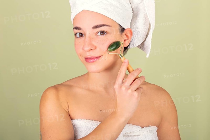 A woman with a towel wrap uses a jade roller for facial massage, promoting skin care and relaxation. Her serene expression reflects self-care and wellness