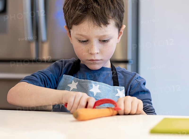 A focused young child wearing a star patterned apron peels a carrot in a bright kitchen environment. The image captures concentration and learning in a domestic setting