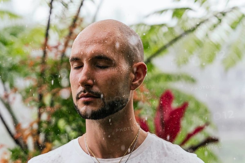 A man stands with closed eyes, embracing the gentle rainfall. Surrounded by lush greenery, he embodies mindfulness, tranquility, and a deep connection with nature