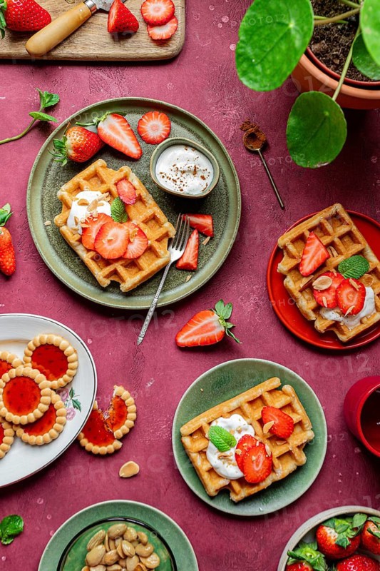 A rustic breakfast scene with homemade waffles topped with fresh strawberries and creamy yoghurt. The setting is adorned with mint leaves and almonds for a sweet, romantic touch