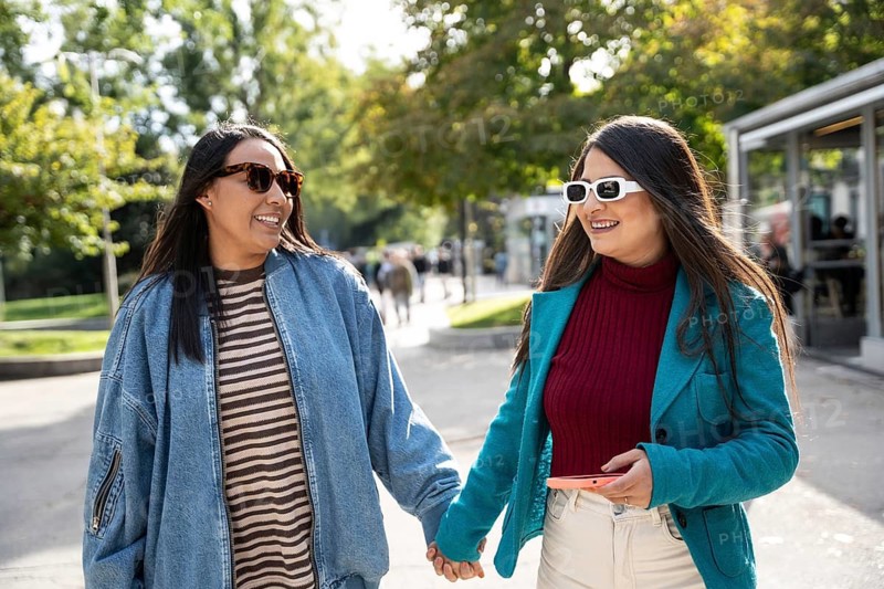 A joyful Latin lesbian couple strolls hand in hand through a lush, sunlit park. Both are wearing sunglasses and casual attire, sharing smiles and happy moments