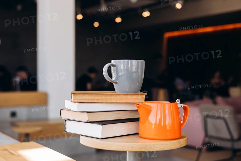 A cozy cafe scene featuring a stack of books on a wooden table, accompanied by a gray mug and a vibrant orange teapot. The blurred background adds a warm ambiance