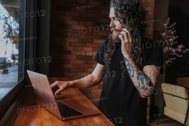 A bearded man with long hair and tattoos uses a laptop while talking on the phone, positioned near a large window in a cozy cafe setting. Natural light enhances the scene