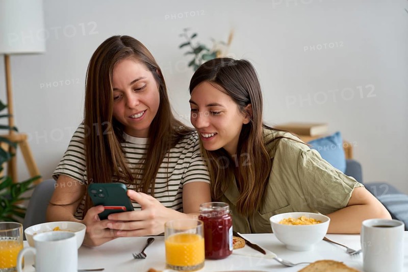 A lesbian couple shares a warm moment at breakfast, smiling as they look at a phone together The setting is cozy, with breakfast items on the table LGBTQIA+ pride