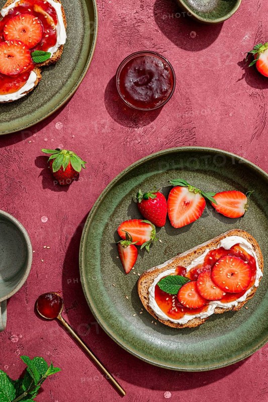 Top view of a delicious breakfast featuring toast topped with cream cheese and fresh strawberries on green plates. A spoon and jam complete the vibrant setup
