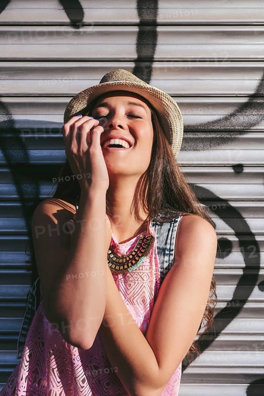 A cheerful woman in a hat and pink top enjoys a sunny day, standing against a metal shutter background. Her joyful expression radiates free-spirited energy in an urban setting