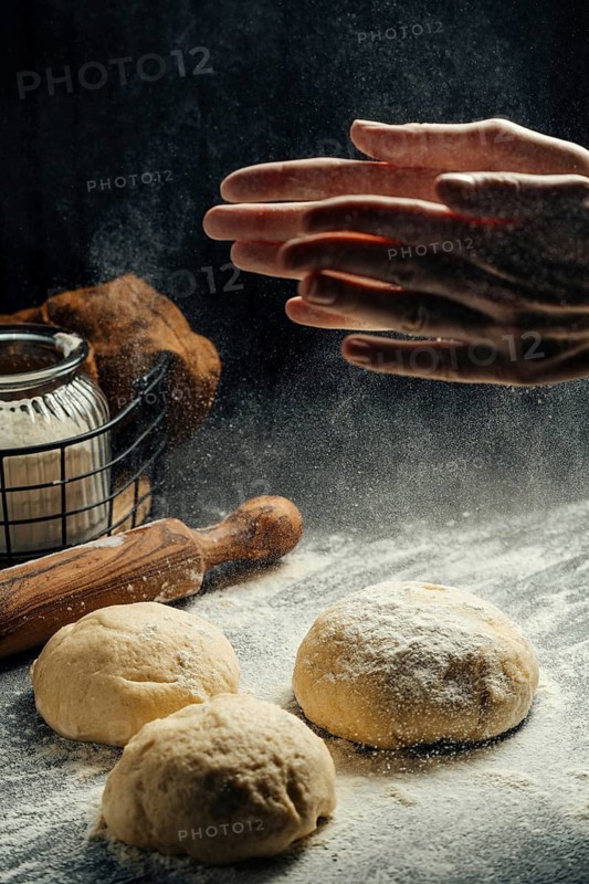A pair of hands dusts flour over dough balls in a rustic kitchen, ready for baking. A rolling pin, and jar are visible, evoking a warm, homey atmosphere