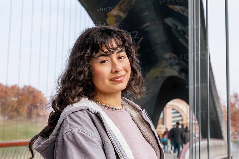 A young Latin woman with curly hair and a relaxed smile, casually dressed, stands confidently on a modern bridge The urban backdrop adds a vibrant touch to the scene