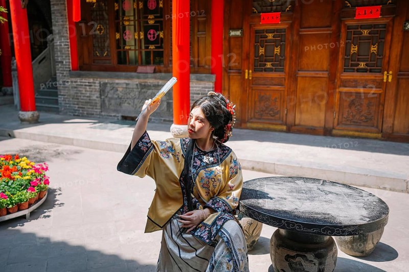 A young woman in traditional Qing Dynasty attire gracefully sits outdoors in Pingyao, China, while shading herself with an elegant fan under the bright sun