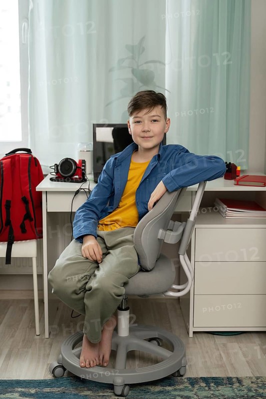 A young boy sits comfortably on a chair in a modern study room with a desk, computer, and red backpack. The space is well-lit and organized, creating a peaceful ambiance