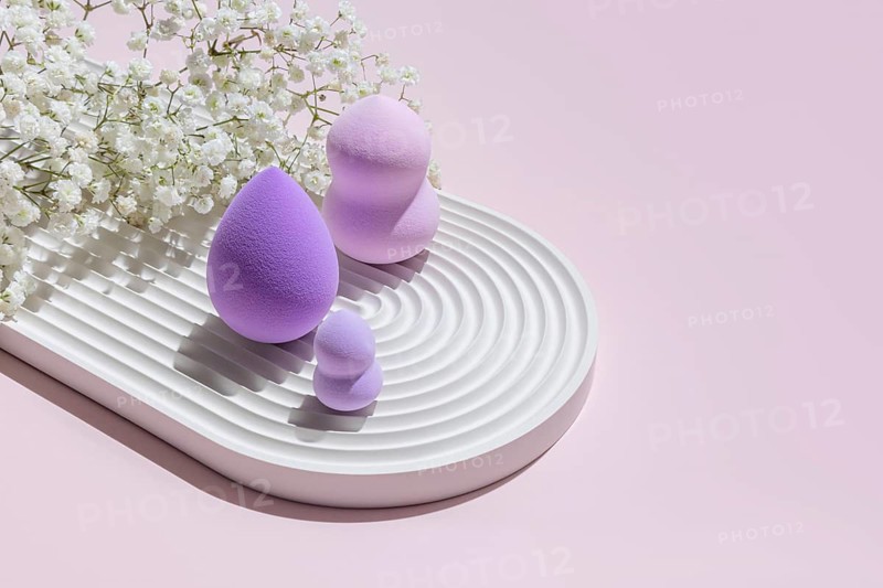 Various sizes of purple beauty blender makeup sponges arranged artistically on a white textured plate, complemented by delicate white flowers on a soft pink background