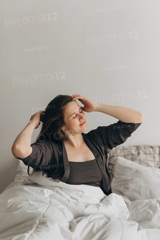A woman in casual wear sits comfortably on a bed, savoring a serene morning moment. Her relaxed pose reflects tranquility, making it a perfect theme for wellness and lifestyle content