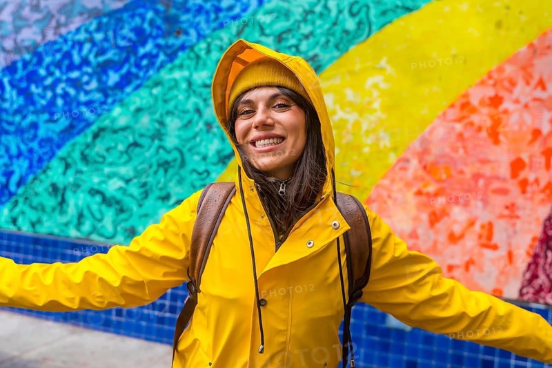 Young woman in a yellow raincoat and beanie spreads her arms, smiling with a backpack against a vibrant rainbow mosaic wall in san francisco, joyful urban exploration