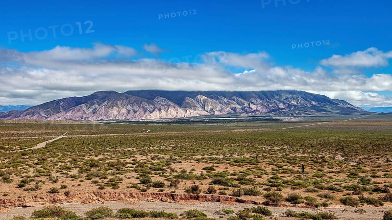 Wide plain in front of a mountain range under a blue, cloudy sky, the landscape and colorful rock formations of the Quebrada de Cafayate near Salta in Argentina