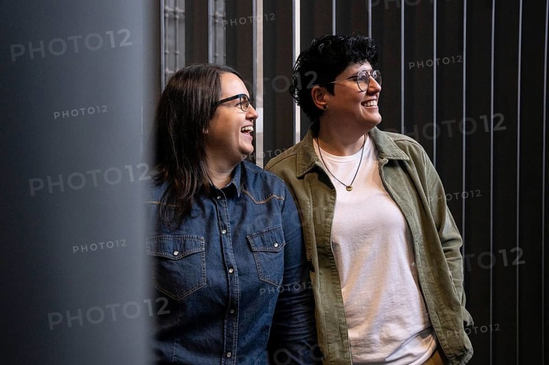 A lesbian couple enjoys a moment of laughter together. Both wear casual attire and glasses, standing against a modern backdrop with a warm, vibe