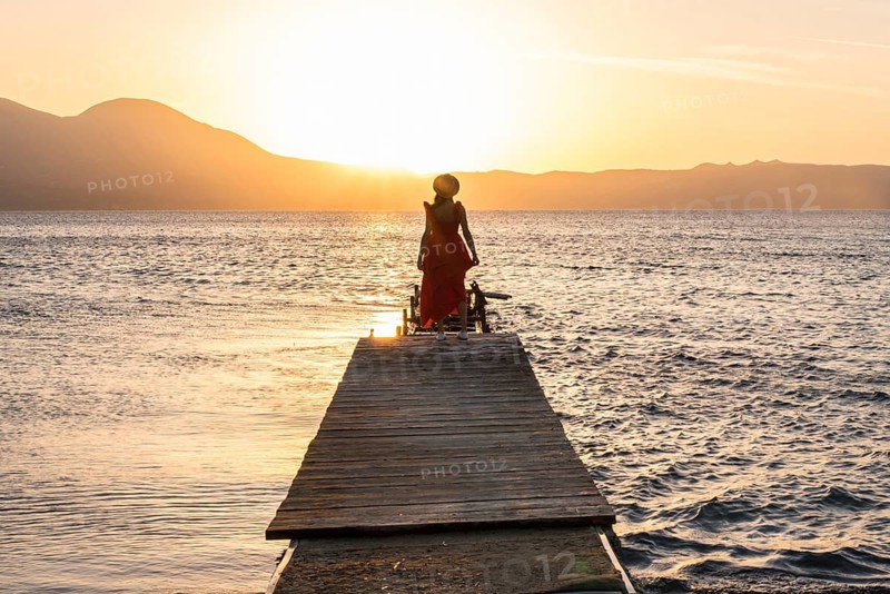 A serene sunset at Papikinou Beach, Milos Island, Greece. A woman in silhouette stands on a pier overlooking calm waters with mountains in the background