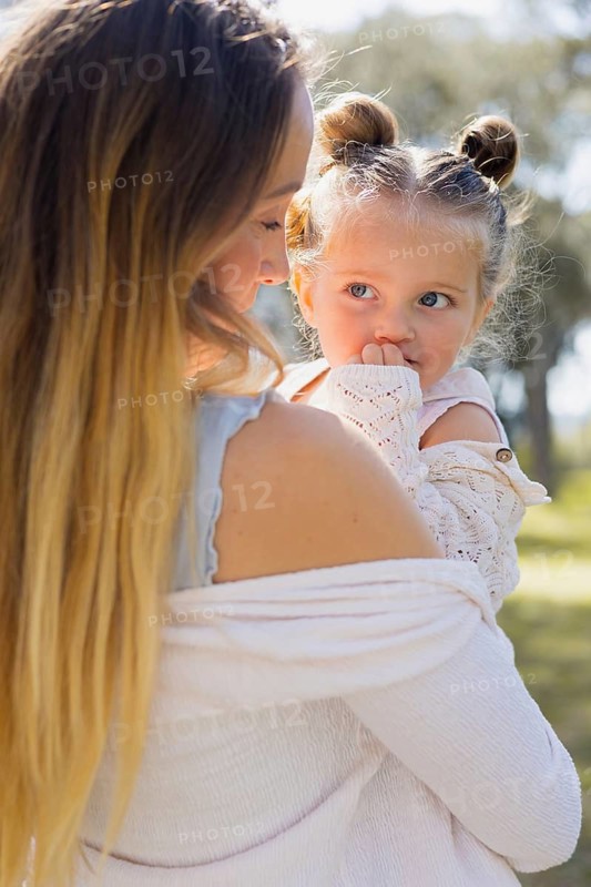 A pregnant woman lovingly holds her young daughter on a sunny day outside. They share a tender moment in a natural setting, highlighting family bonding and affection