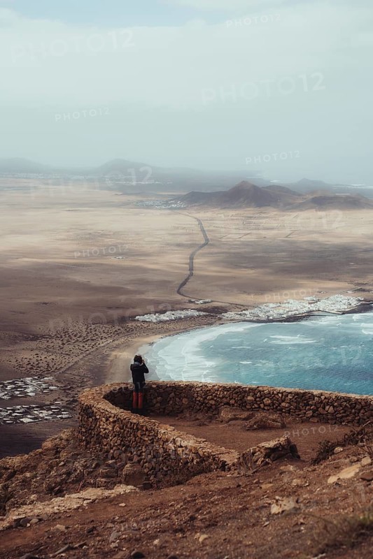 A stunning view of Lanzarote's coastline, showcasing the contrast between the blue ocean and arid volcanic terrain. A person stands at a stone wall, capturing the scene