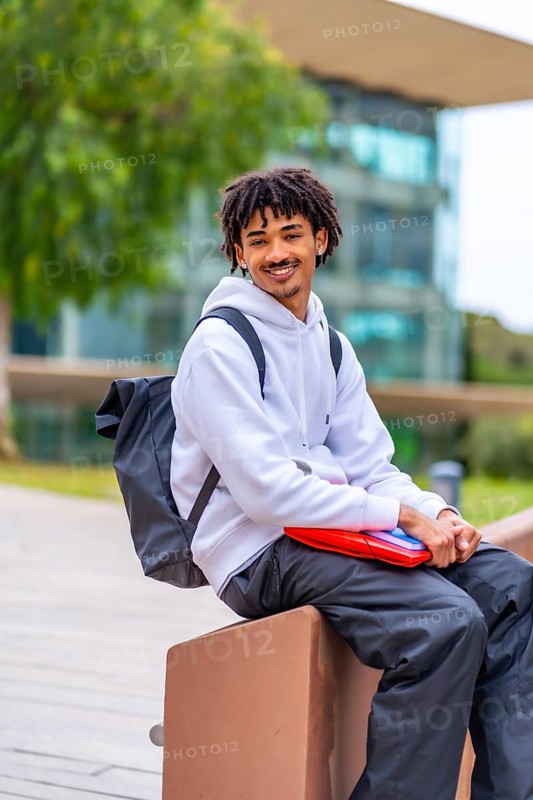 Vertical side view of a cool smiling african american student with dreadlocks waiting sitting outside the university
