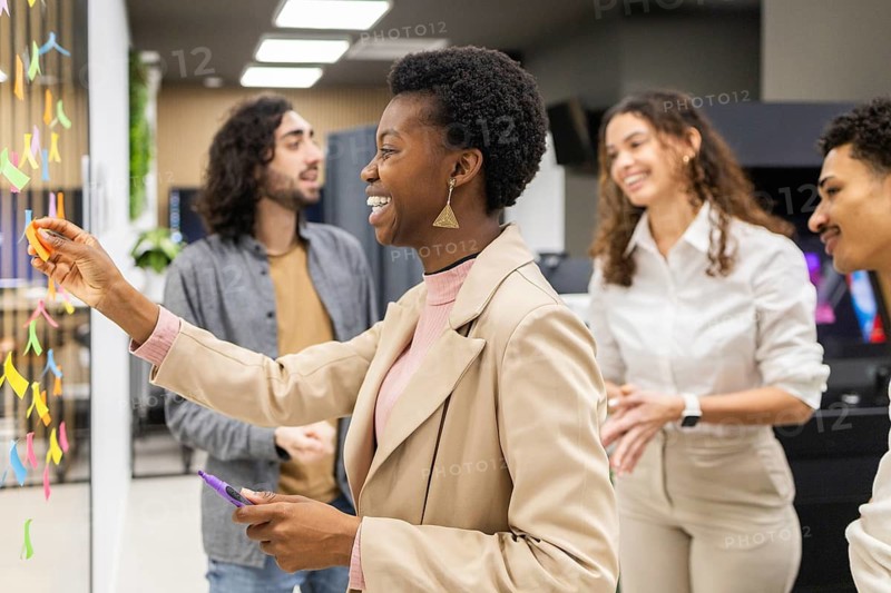 A diverse group of colleagues engaged in a brainstorming session in a modern office. Smiling and interacting, they are using colorful sticky notes on a glass wall